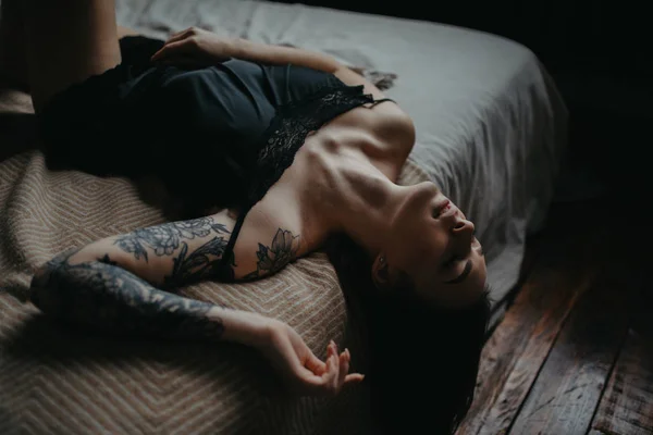 Mujer joven en lencería negra con tatuaje en el brazo se encuentra en la cama —  Fotos de Stock