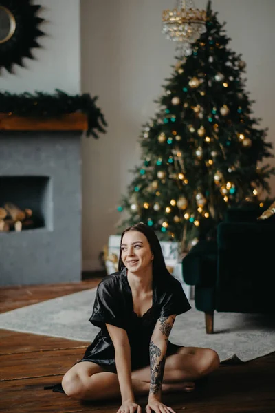 Mujer joven con tatuaje en el brazo se sienta al lado del árbol de Navidad . — Foto de Stock