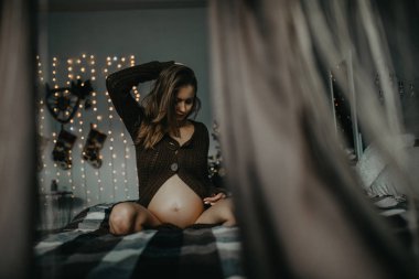 Hamile kadın, Noel süslemelerinin arka planında yatakta oturuyor..