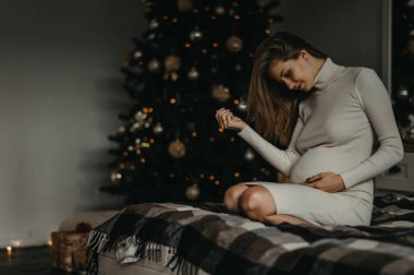 Hamile kadın, Noel ağacının arka planında yatakta oturuyor..