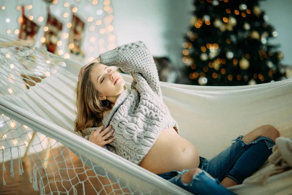 Έγκυος γυναίκα βρίσκεται και στηρίζεται σε αιώρα στο φόντο του χριστουγεννιάτικου δέντρου . — Φωτογραφία Αρχείου