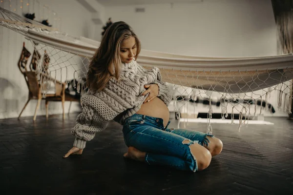 Těhotná žena ve svetru a džíny sedí na podlaze. — Stock fotografie