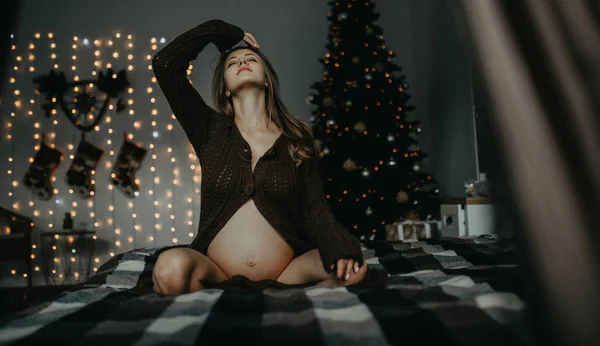 Kobieta w ciąży siedzi na łóżku na tle dekoracji świątecznych. — Zdjęcie stockowe