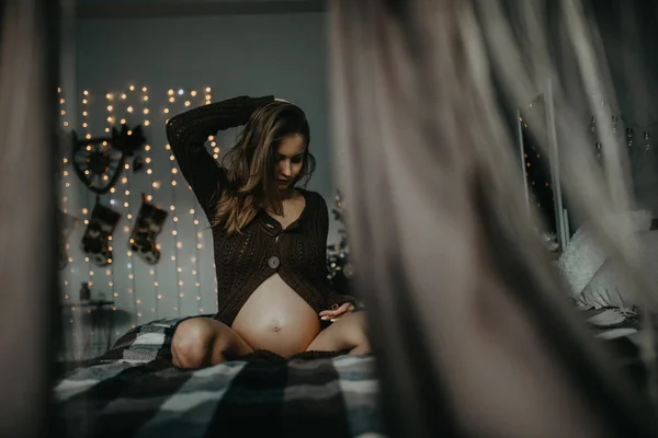 Pregnant woman sits on bed against background of Christmas decorations. — Stock Photo, Image