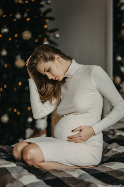 Έγκυος γυναίκα κάθεται στο κρεβάτι με φόντο το χριστουγεννιάτικο δέντρο. — Φωτογραφία Αρχείου