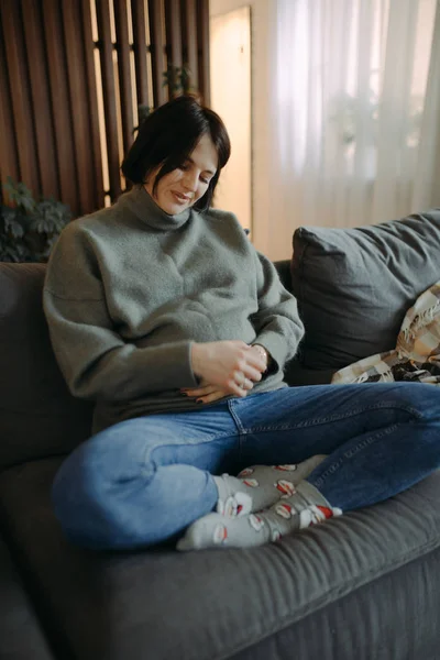 Těhotná žena sedí na pohovce a dotýká se svého břicha. — Stock fotografie
