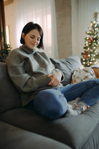 妊娠中の女性は座っていると彼女のおなかに触れますクリスマスツリーの背景に. — ストック写真