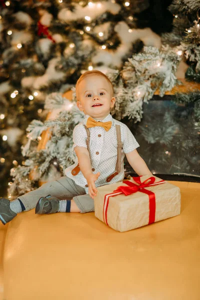 Un niño pequeño se sienta en la capucha de un coche con caja de regalo sobre el fondo de un árbol de Navidad —  Fotos de Stock
