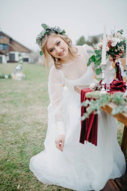 Gelin şamdanlar, tabaklar, şarap kadehleri, çiçek aranjmanı ve kırmızı bir masa örtüsüyle ziyafet masasının yanına oturur..