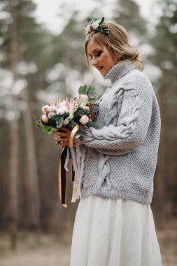 Gelin ormanda beyaz elbise ve kazak giyerek duruyor. Yakın çekim..