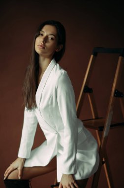 Beyaz ceketli ve çizmeli genç bir kadın stüdyoda merdivenin yanında poz veriyor..