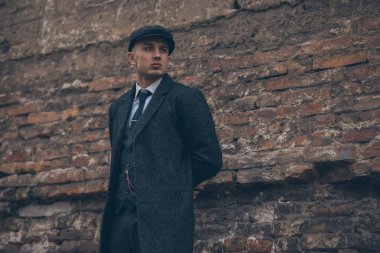 1920 'lerde Peaky at gözlüğü takmış eski tuğla duvarın yanında eski bir İngiliz gangsteri gibi poz veren bir adam..