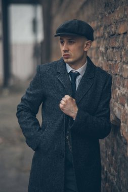 A man posing in the image of an English retro gangster of the 1920s dressed in Peaky blinders style near old brick wall. clipart