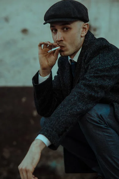 A man smokes a cigarette in the image of an English retro gangster of the 1920s dressed in a coat, suit and flat cap in Peaky blinders style.