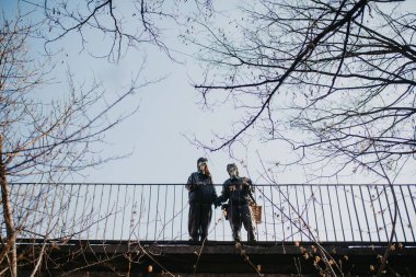 Birbirine aşık bir çift köprüde yürüyor ve NBC koruyucu giysileri ve gaz maskeleriyle el ele tutuşuyor. Koronavirüs COVID için önleyici önlemler ve koruma kavramı 19 salgın hastalık ve diğer küresel tehlikeler.