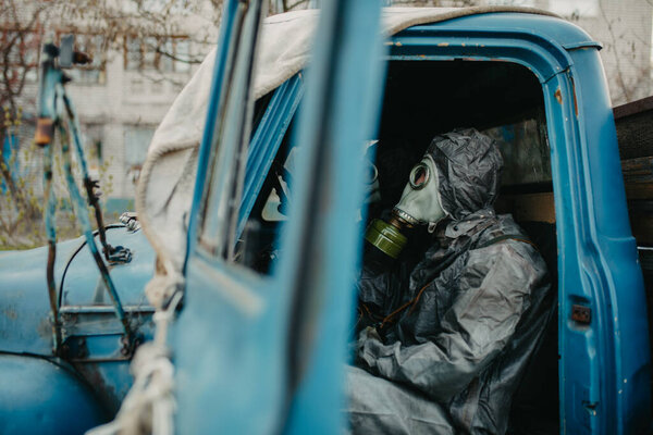Two persons sits in NBC protective suits and gas masks in old truck. Concept of a preventive measures and protection for coronavirus COVID 19 pandemic and other global dangers.
