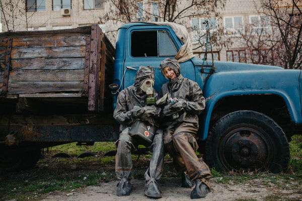 Couple in love sits in NBC protective suits and gas mask near old truck. Concept of a preventive measures and protection for coronavirus COVID 19 pandemic and other global dangers.