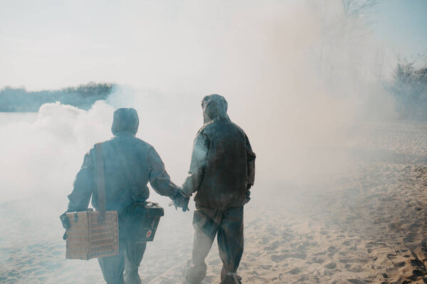 Couple in love walks in NBC protective suits and gas masks on smoke background. Concept of a preventive measures and protection for apocalypse, global pollutions of environment and pandemic.