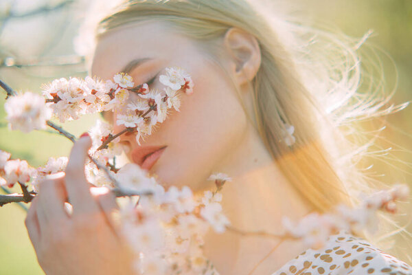 Portrait of young blonde woman among blossoming branches of apricot trees in spring garden.