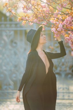 Siyah elbiseli ve şapkalı genç kadın sakura ağaçlarıyla parkta rahatlıyor ve eğleniyor..