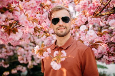 Güneş gözlüklü genç adamın portresi. Parkta sakura ağaçlarıyla eğleniyor ve dinleniyor..