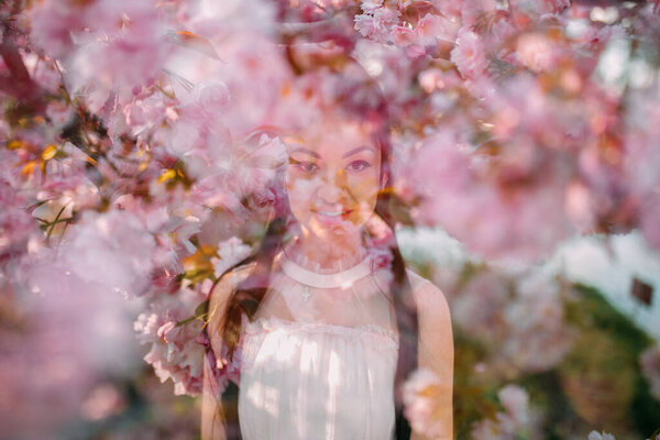 Portrait of young woman in white dress in park among blooming sakura trees with effect of multiple exposure.