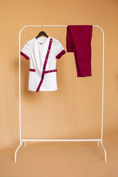 White blouse and burgundy pants for medical workers are on a rack hanger on an orange background.