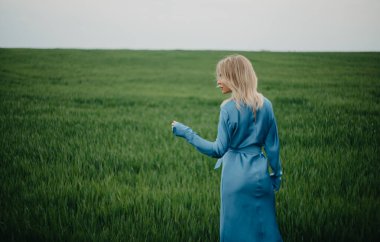 Mavi elbiseli genç bir kadın yeşil buğday tarlalarının arasında yürüyor. Arkadan bak. Boşluğu kopyala.