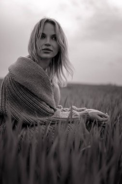 Ceketli genç bir kadın buğday tarlasında oturuyor. Siyah ve beyaz resim.