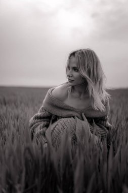 Ceketli genç bir kadın buğday tarlasında oturuyor. Siyah ve beyaz resim.