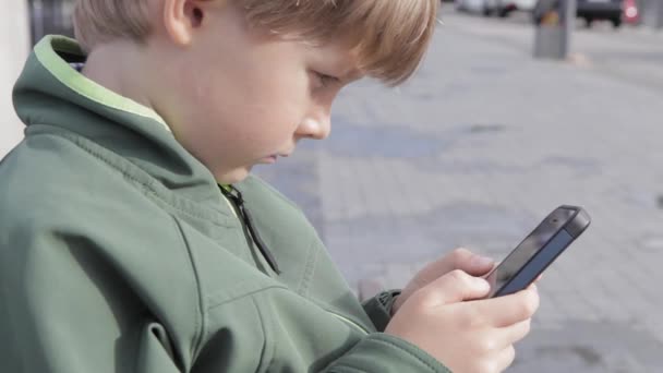 Młody chłopak grać na smartfonie w mieście. Chłopiec z telefonu komórkowego. Mały chłopiec na ulicy gry z smartphone — Wideo stockowe