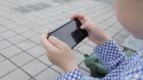Νεαρό αγόρι παίζει στο smartphone στην πόλη. Αγόρι με κινητό τηλέφωνο. Μικρό αγόρι στο δρόμο παίζοντας με smartphone — Αρχείο Βίντεο