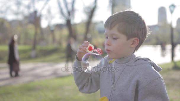 Un niño rubio juega en la calle. Sopla burbujas. Chico joven soplando burbujas, cámara lenta . — Vídeos de Stock