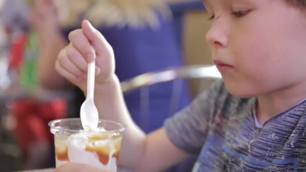 Um rapazinho está a comer gelado num café. O menino loiro no café está comendo sorvete com uma colher . — Vídeo de Stock