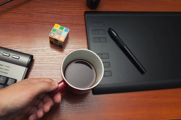 Pauză, odihnă în timpul procesului de lucru, o ceașcă de cafea, un cub de rubik, un puzzle. Tabletă grafică pe o masă din lemn — Fotografie, imagine de stoc