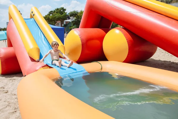 El chico se entretiene al aire libre, bañado en una piscina inflable. Desliza hacia abajo desde el tobogán en el agua . Imagen De Stock