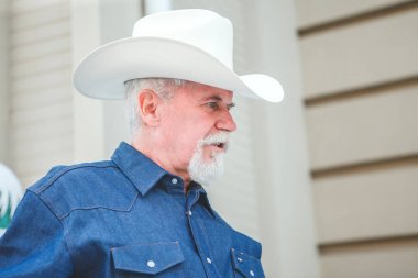 Olgun bir kovboy şapkası, kot pantolon ve bir kot gömlek kameraya benziyor. Açık hava.