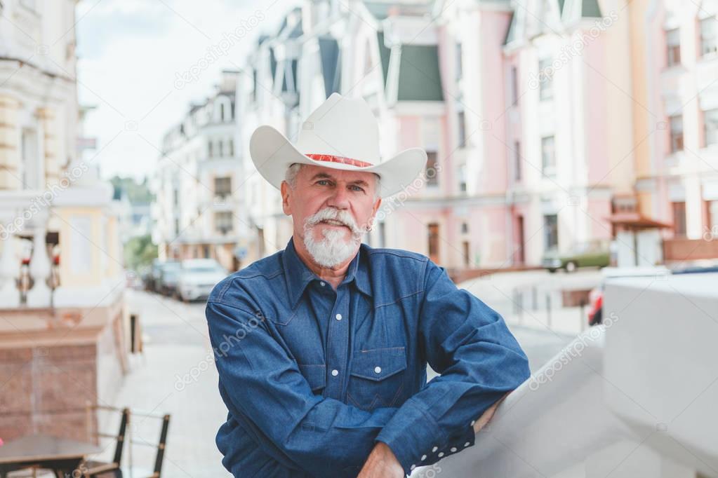 A mature cowboy in a hat, jeans and a denim shirt looks at the camera. On open air.