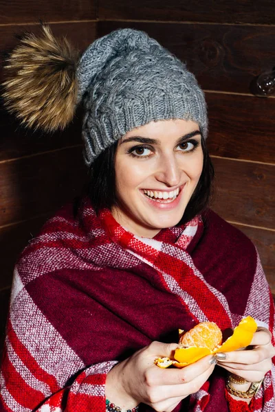 Настроение зимы. Молодая красивая темноволосая женщина улыбается в одежде и кепке с мандаринами на деревянном фоне . — стоковое фото