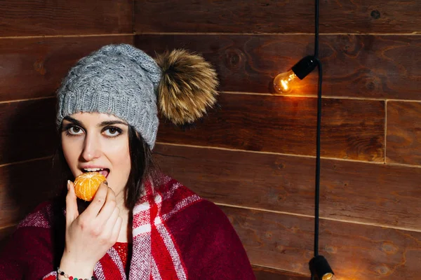 Молодая красивая темноволосая женщина улыбается в зимней одежде и кепке с мандаринами на деревянном фоне . — стоковое фото