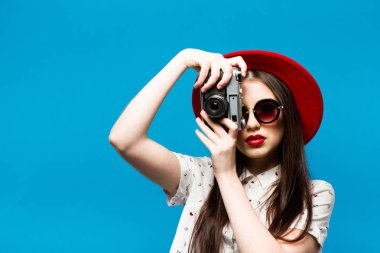 Moda güzel genç gülümseyen kadın retro kamera kırmızı şapka, mavi arka plan üzerinde tutar.