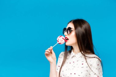 Üzerinde mavi arka plan izole lolipop tutan genç güzel kadın. Mutlu kız çok yeme güneş gözlüğü takıyor şeker renkli. Eğleniyor lolipop yemek eğlenceli ve neşeli kadın.