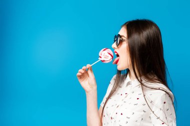 Üzerinde mavi arka plan izole lolipop tutan genç güzel kadın. Mutlu kız çok yeme güneş gözlüğü takıyor şeker renkli. Eğleniyor lolipop yemek eğlenceli ve neşeli kadın.