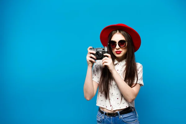 Moda bella giovane donna sorridente tiene fotocamera retrò indossando cappello rosso, su sfondo blu . — Foto Stock
