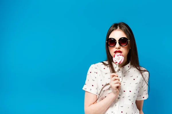 Mujer hermosa joven sosteniendo piruleta aislado sobre fondo azul. Chica feliz con gafas de sol comiendo caramelos multicolores. alegre y alegre mujer comer piruleta divertirse . — Foto de Stock