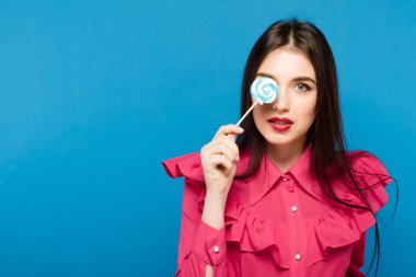 Genç kadın yüzünü lolipop ile mavi arka plan üzerinde kaplayan. Gömlek ve pantolon giyiyor. Kameraya bakarak