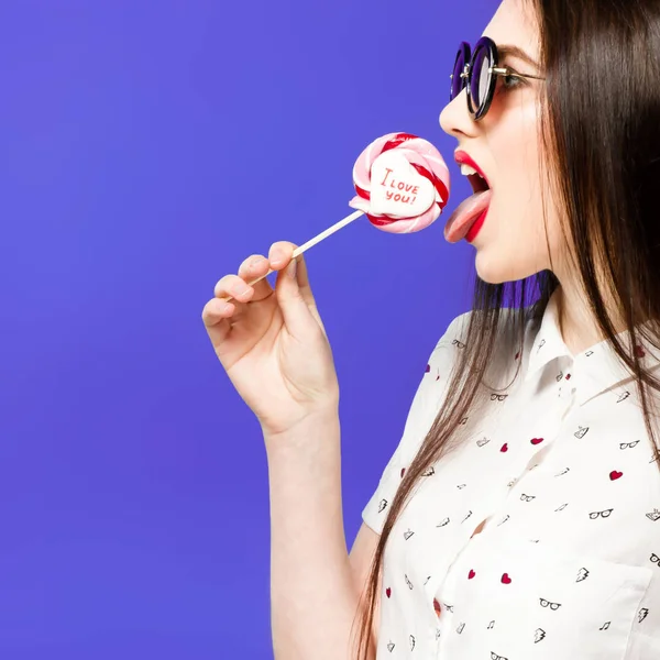 Jeune belle femme tenant sucette isolée sur fond bleu. Fille heureuse portant des lunettes de soleil manger des bonbons multicolores. joyeuse et gaie femme manger sucette avoir du plaisir — Photo