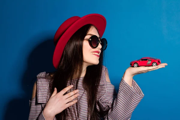 Schönes Modell in modischen roten Hut und eine rote Kupplung mit Auto auf blauem Hintergrund — Stockfoto