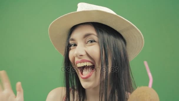 Молода дівчина в купальнику і літньому капелюсі п'є кокосовий коктейль і посміхається. Зелений фон — стокове відео