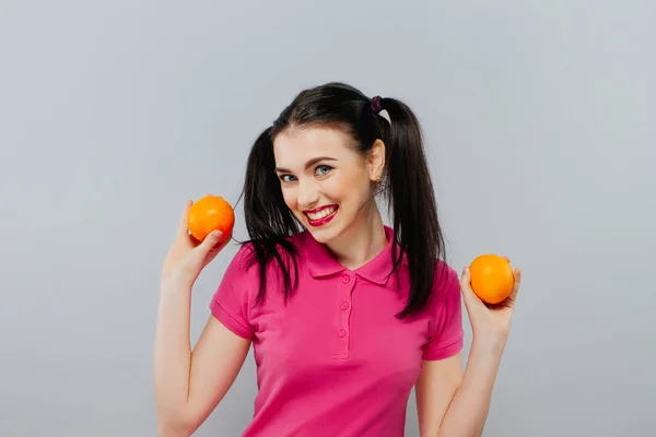 Atractiva joven sonriente posando con dos naranjas frescas sobre fondo gris . — Foto de Stock